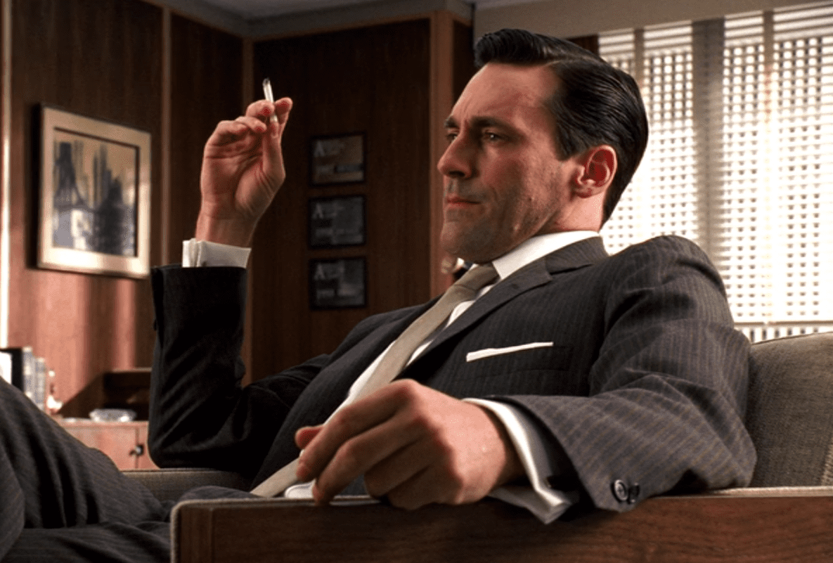 Don Draper sits in a chair smoking a cigarette