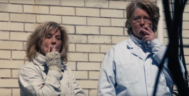 Gladys and another member of the Guilty Remnant, dressed in white and smoking in front of a brick wall