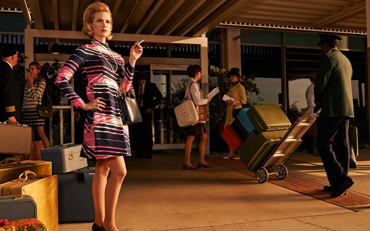 betty draper smokes a cigarette while standing by hoards of luggage at a posh hotel