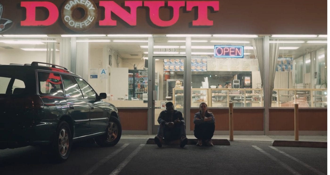 a male and female couple sit on the floor of a parking lot outside a donut shop