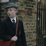 a young english schoolboy in his school uniform with red school bag standing next to a brick wall and wrought iron fence simon burrows the romanoffs