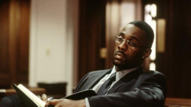 Stringer Bell (Idris Elba) sits in a courtroom wearing glasses and holding a book in The Wire's pilot episode