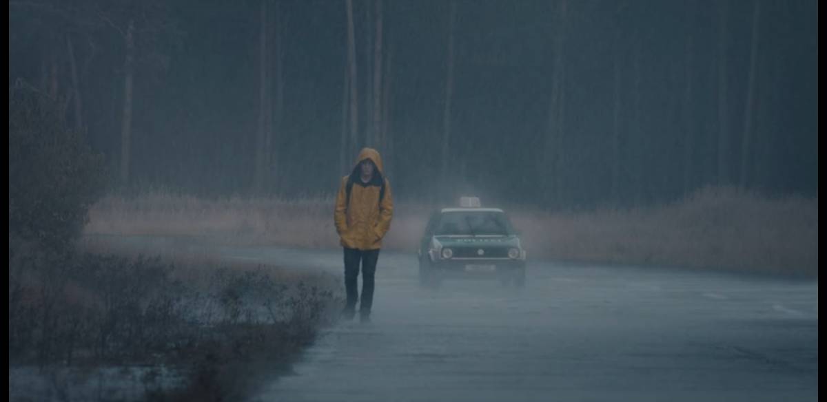 Jonas Kahnwald walking along the road in the rain next to a police car in Netflix's Dark