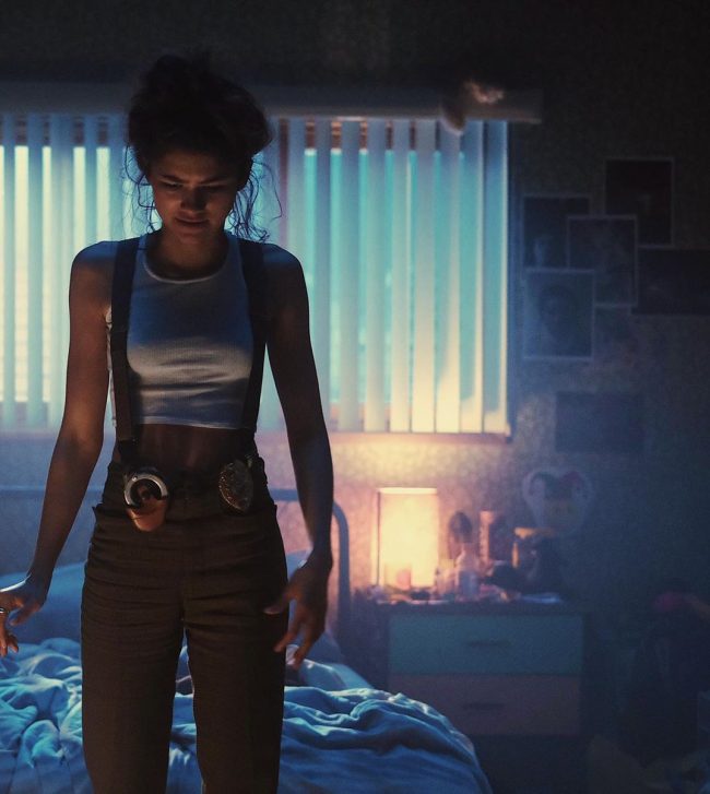 Rue standing in her bedroom while wearing a gun holster, handcuffs, and police badge on her hip.