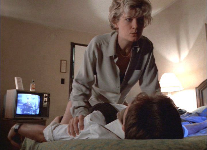 Detective White aggressively straddles Agent Mulder on the bed in his hotel room.