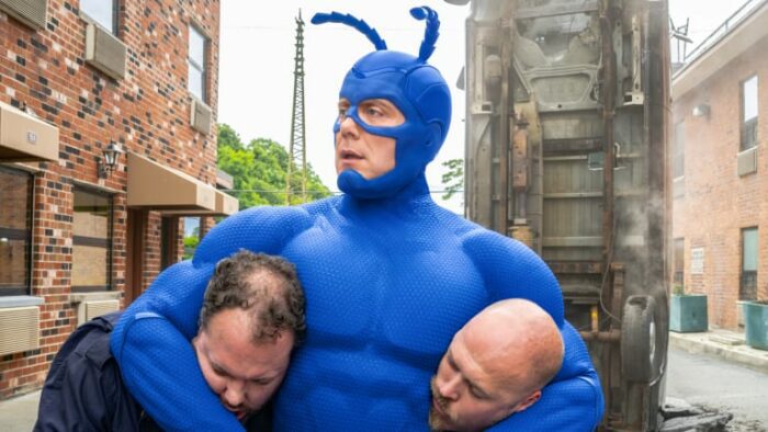 The Tick (‘16) with two criminals in headlocks with an overturned car in the background 