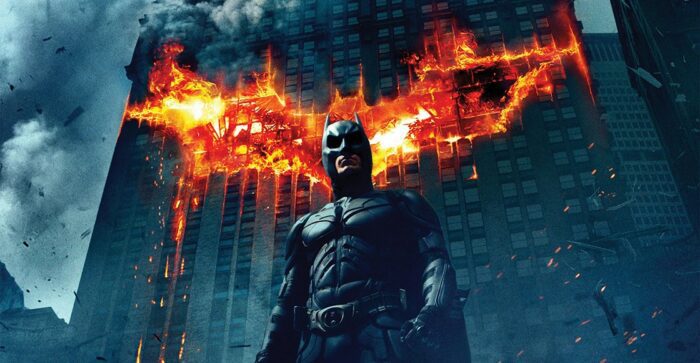 Batman stands in front of a building which has a flaming bat upon it 