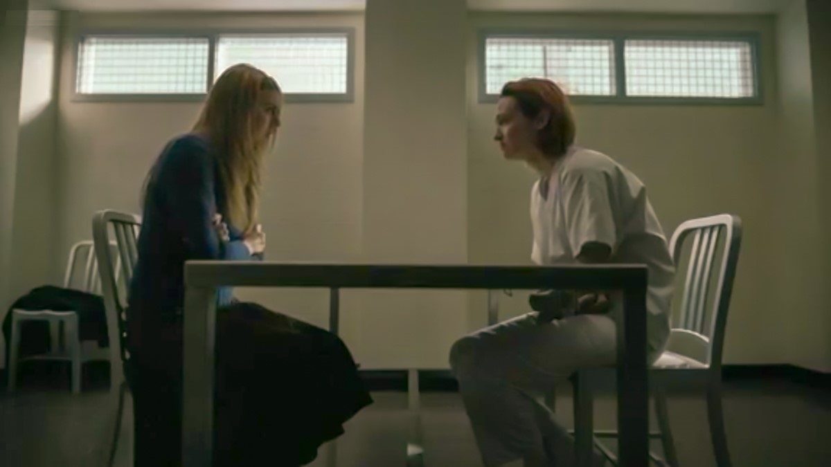 Holly and Lou sit, facing each other, in a stark white hospital room, Holly looks uncomfortable.