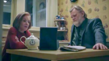 Ira and Bill sit having tea in her kitchen at the dining room table, looking at a photo frame