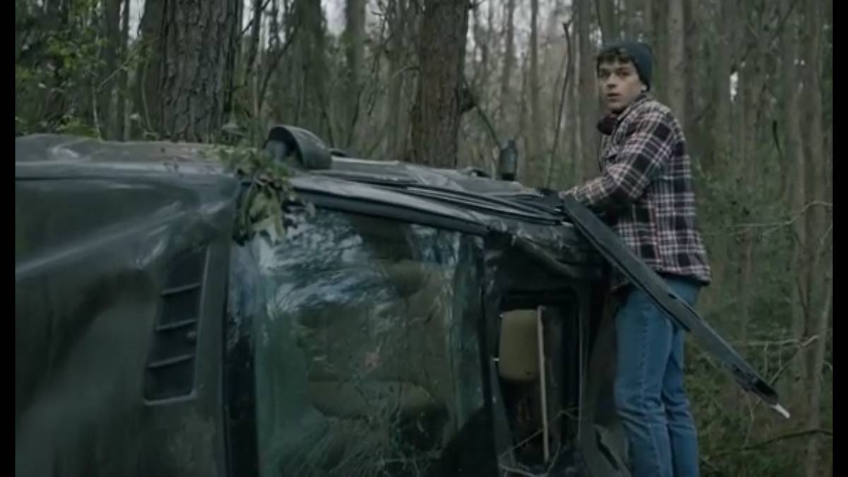 Pete stands on the side of the wreckage of a car in the woods, looking around.