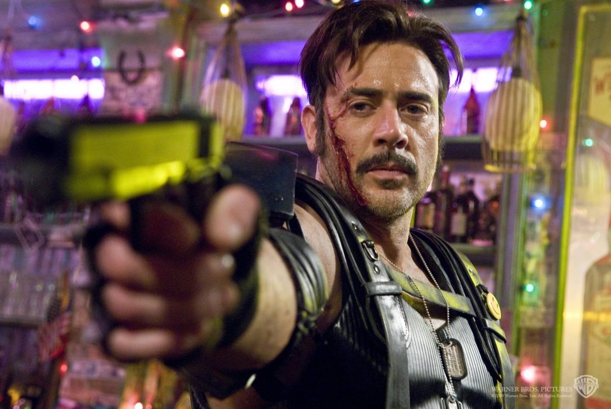 Image from the 2009 Watchmen film depicting the Comedian in a Vietnamese bar with an open bleeding gash on his cheek and jaw, aiming a revolver that he has raised toward the viewer.