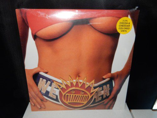 A close up of a woman's torso, breasts falling out of her shirt, and a chamionship belt featureing the name of the band Ween.