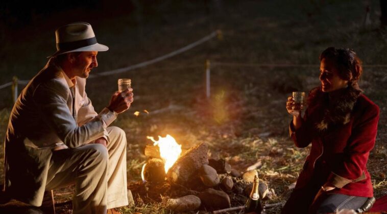 Roma and Remus sit across the campfire from each other smiling and holding jars for glasses