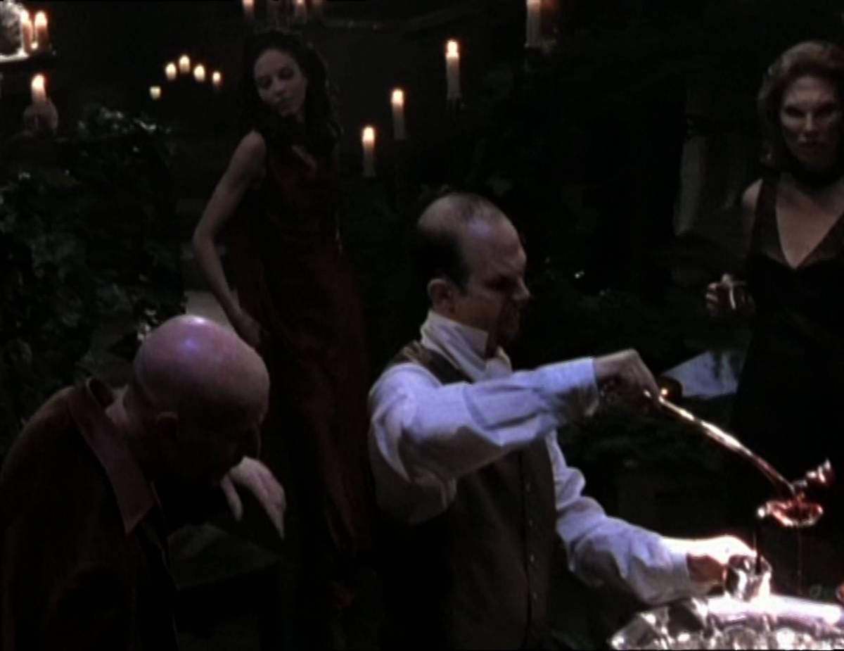 Dru comes downstairs while other vampires get blood from a punch bowl at her party.