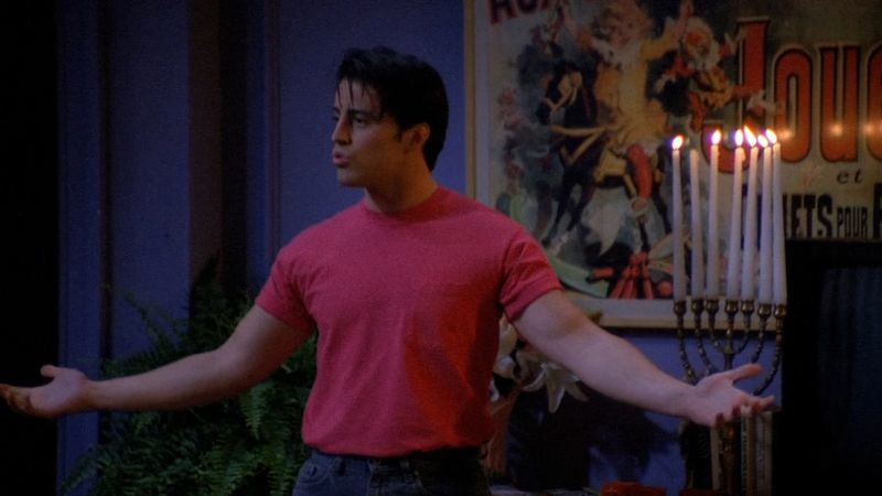 Joey, arms outstretched in a pink t-shirt, delivers an overperformed line on stage in a small theater.
