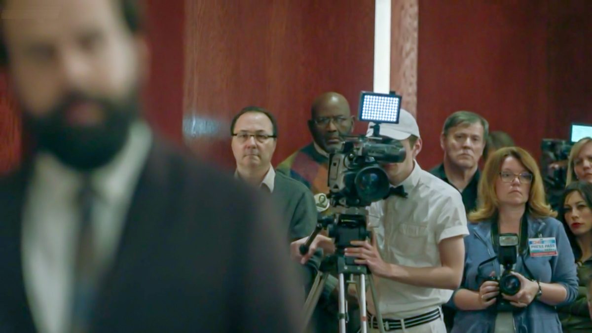 An ice cream man stands behind a TV camera in the courtroom, Finkelstein blury in the foreground. [Mr. Mercedes S03E06]