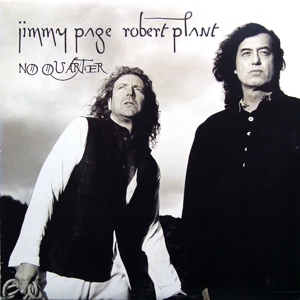 In black and white, we see Robert Plant on the left wearing a black coat with white sleeves, and Jimmy Page stands next to him, all in black.