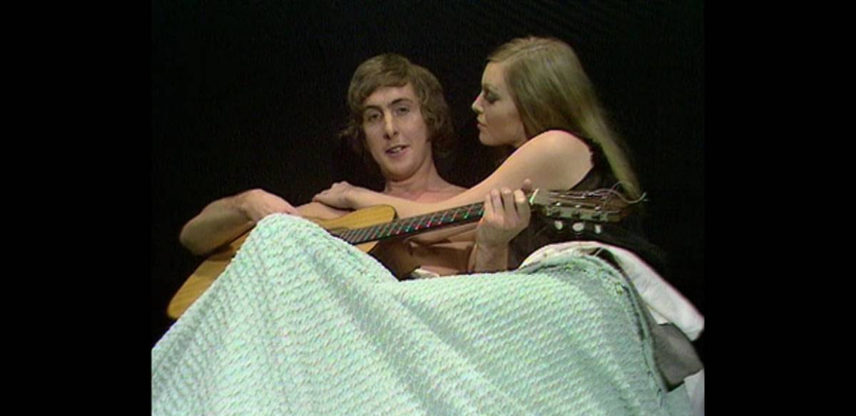 Eric Idle lies in bed playing guitar while being caressed by a beautiful woman