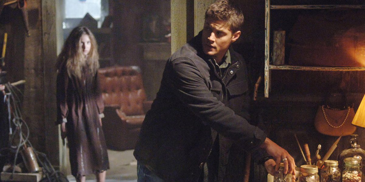 Dean standing, one hand gripping the lid of a jar and looking behind him, creepy little girl lingers in the background watching him