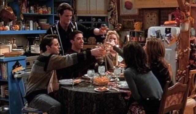 The friends cast sit around a table, toasting wine glasses together after all their Thanksgiving plans had fallen apart.
