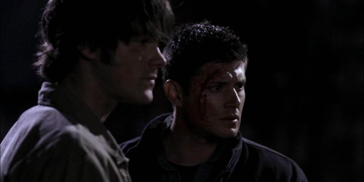 Sam and Dean looking at something in front of them somberly while standing in the dark, with blood from a cut on the left side of Dean's head
