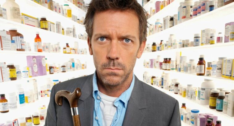 House, looking bemused, stands facing the camera with a corridor of floor to ceiling prescription medication behind him