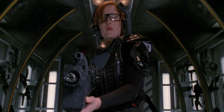 Scully stands in the module that is the entrance to a VR game wearing a VR headset and other game gear and holding a large automatic weapon.