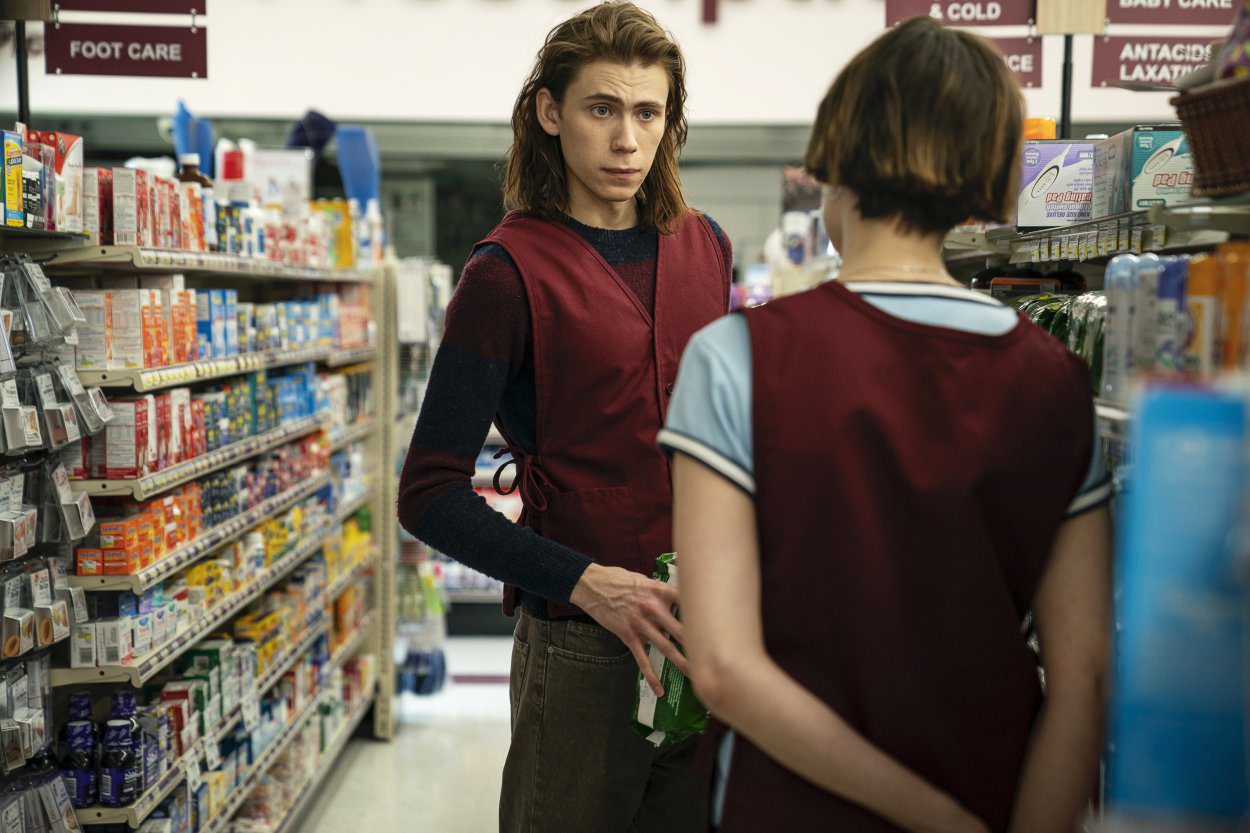 Julian stands in the aisle of the pharmacy talking to his female coworker