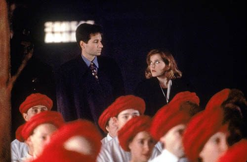 Mulder and Scully look skeptically at one another as they watch from the back of a church full of vergetarions wearing blood red turbins.