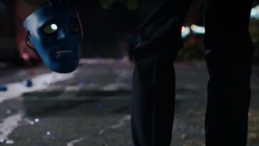 Dr. Manhattan shot from the knees down is shown from behind holding a mask of his likeness.