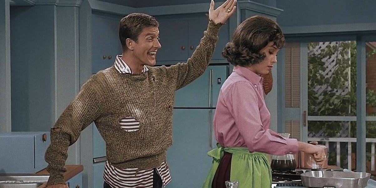 Dick Van Dyke smiling with one arm down and one arm up, Mary Tyler Moore with her back to him cooking eggs, picture is colorized