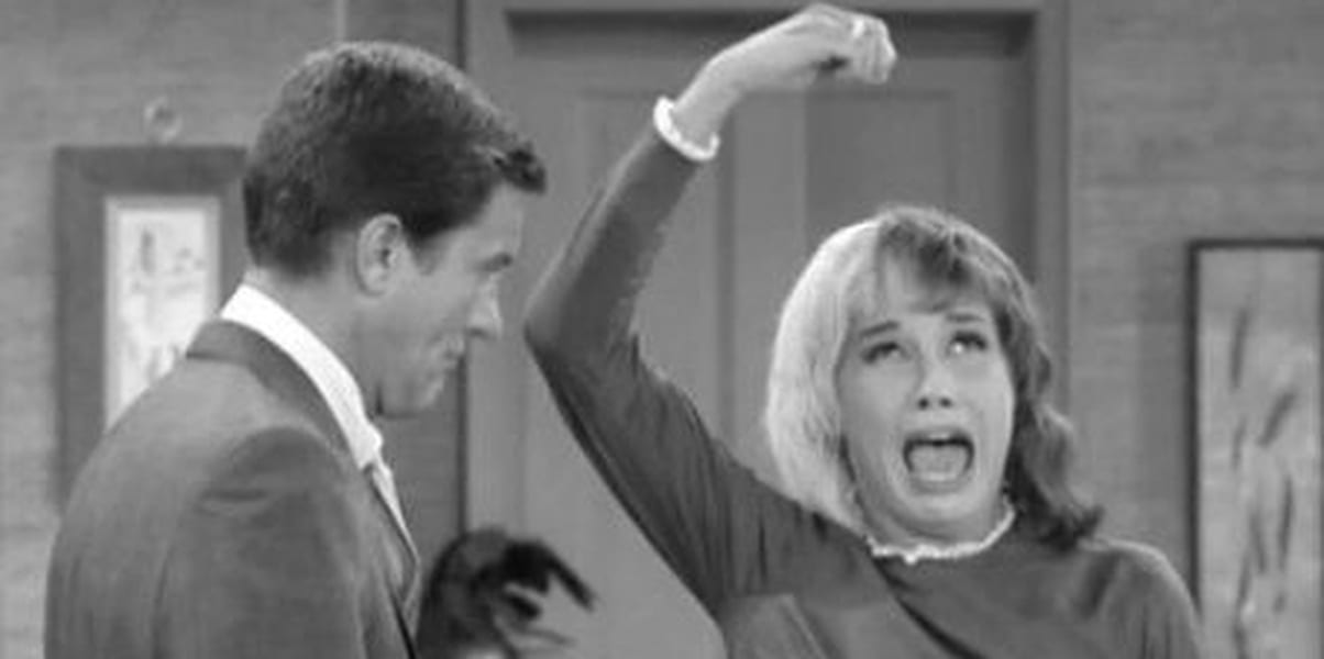 Mary Tyler Moore with an upset look on her face, her arm over her head with her fingers indicating she's holding something, her mouth open, Rob watching her, the image in black and white