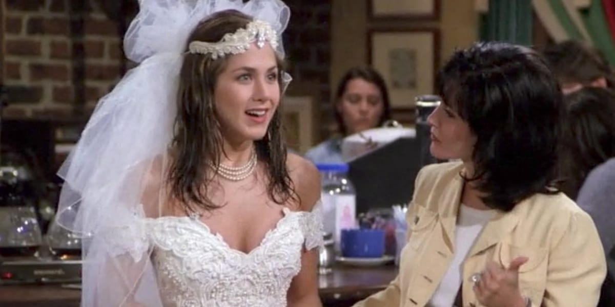 Rachel Green in the Friends pilot wearing a wedding dress with a smile looking ahead with Monica's profile shown looking at Rachel