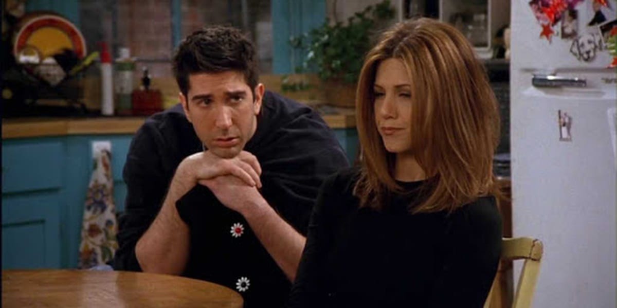 Rachel looking hurt as she stares ahead, Ross sits behind her, hands folds, looking at her sadly but hopefully in Friends