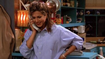 Rachel looking down, smiling, holding a phone to her ear in one hand and her other hand on her hip in Friends