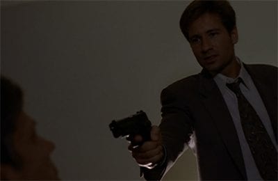Mulder yells and brandishes a gun at CSM, who is sitting in a chair in front of him.