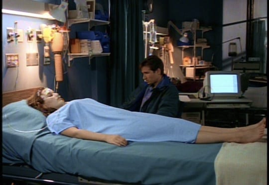 Scully lays on her hospital bed, still in a coma, while Mulder leans towards her and holds her hand.