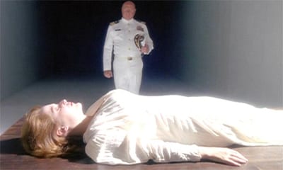 Scully lays flat on a table dressed in white on the bottom half of the frame. Behind her, also in white, is her father, dressed in white and standing straight.