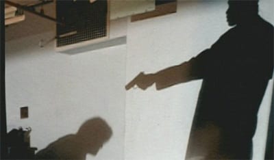 Two shadows, one of a man on the floor. The other is a man above him, pointing a gun directly at him. 
