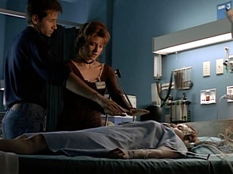 Mulder stands next to Dana Scully’s hospital bed alongside Melissa, who is holding a crystal over Dana