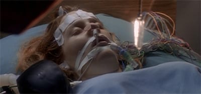 Dana Scully’s head and shoulders are in frame. She’s in a hospital gown, and her face has tape and tubes all over it.