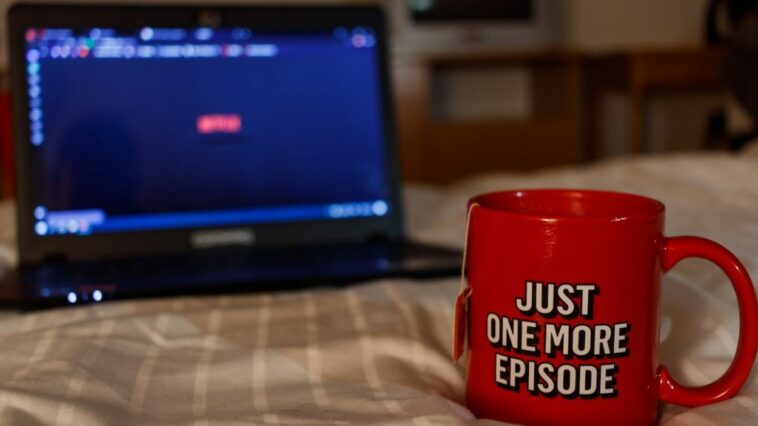 A mug in front of a laptop with the text "Just one more episode" on it.
