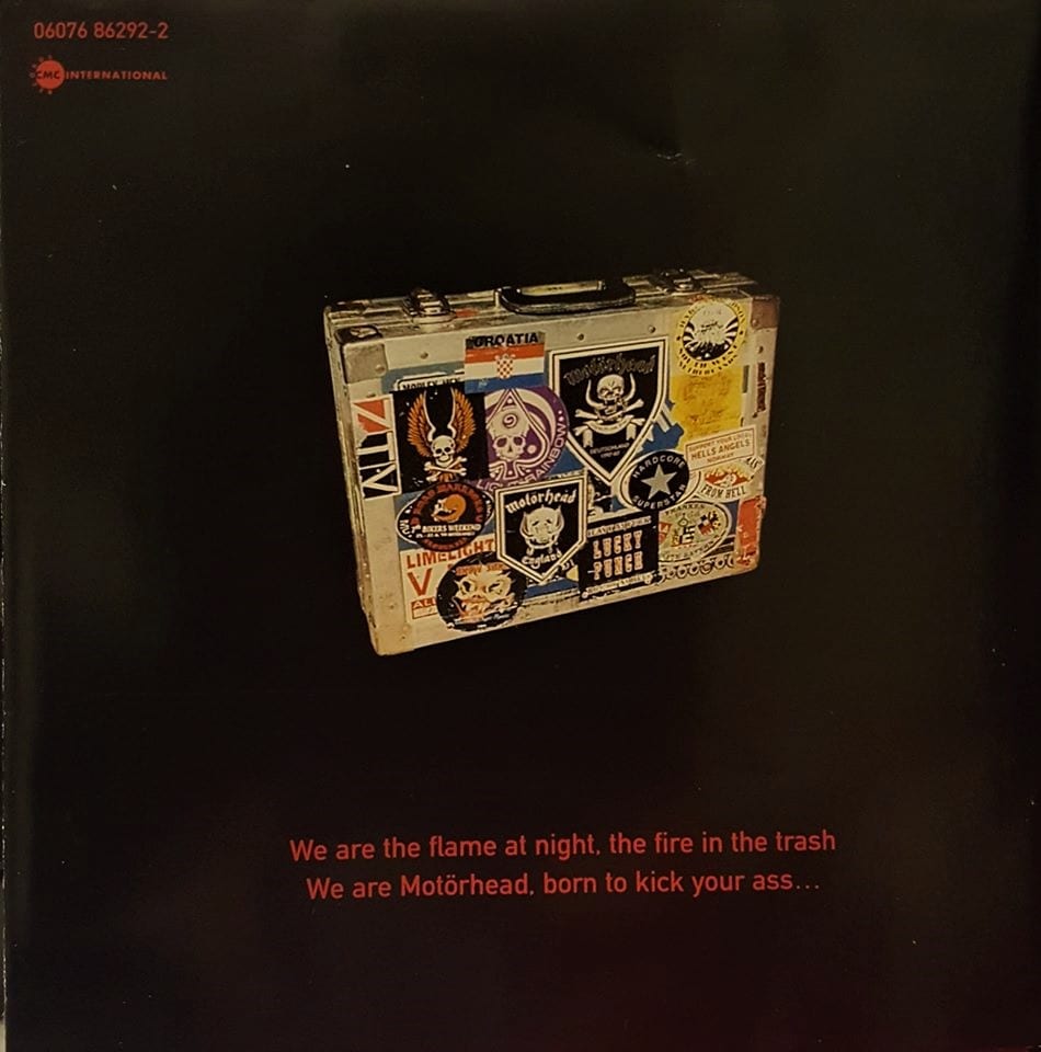 A metal suitcase adorned with various Motorhead and other rock n' roll stickers, stands upright amidst a black background, with a quote from the song "We Are Motorhead" underneath in red letters.