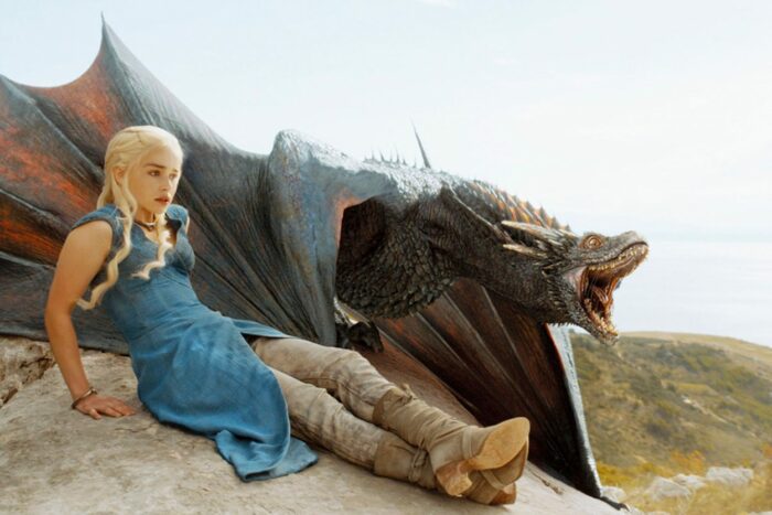 Daenerys Targaryen sits on a rock next to her roaring dragon