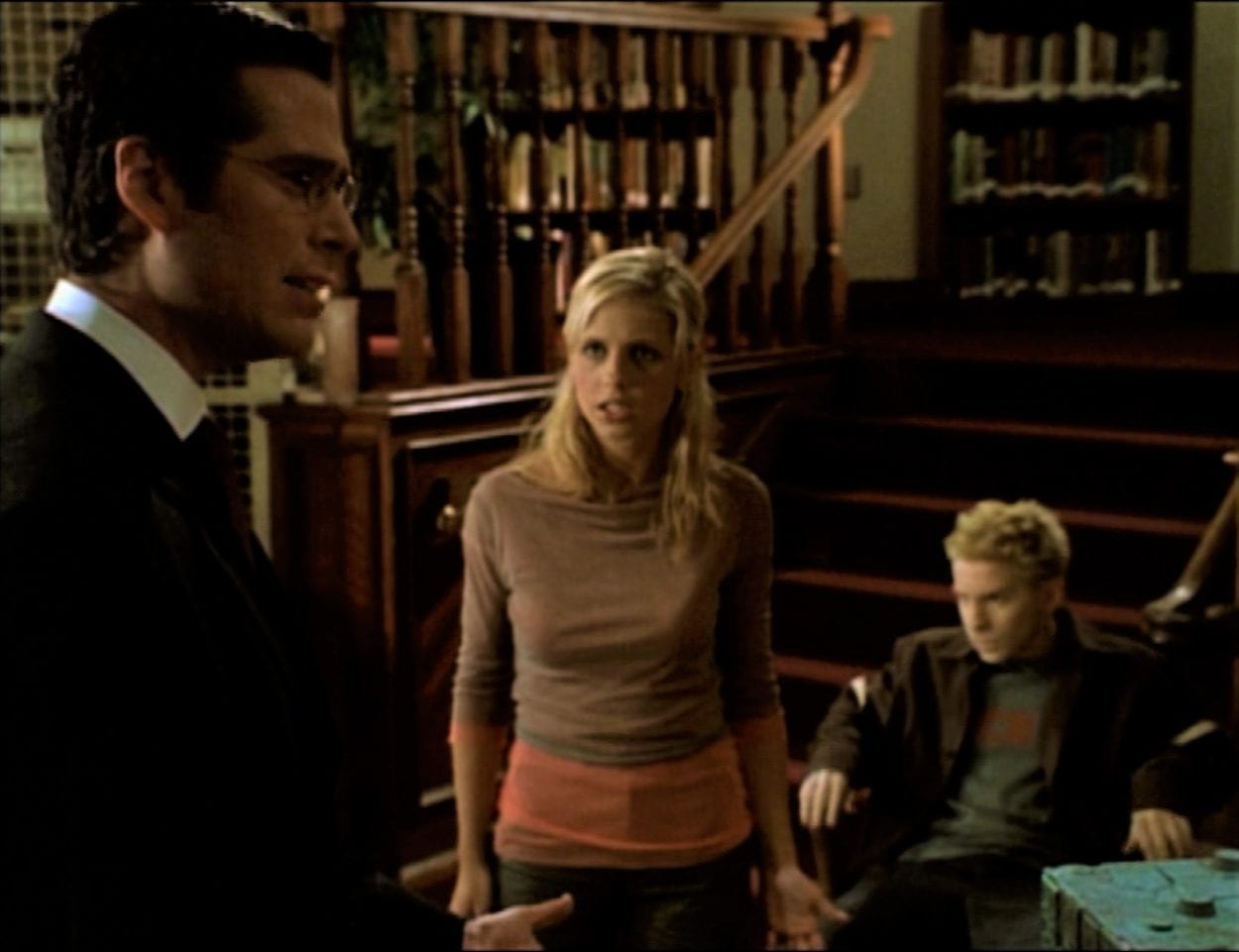 Wesley and Buffy are arguing in the library, while Oz sits in the background