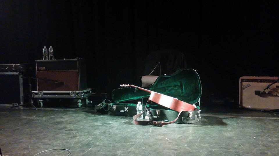 A guitar in its case, on stage and surrounded by amps