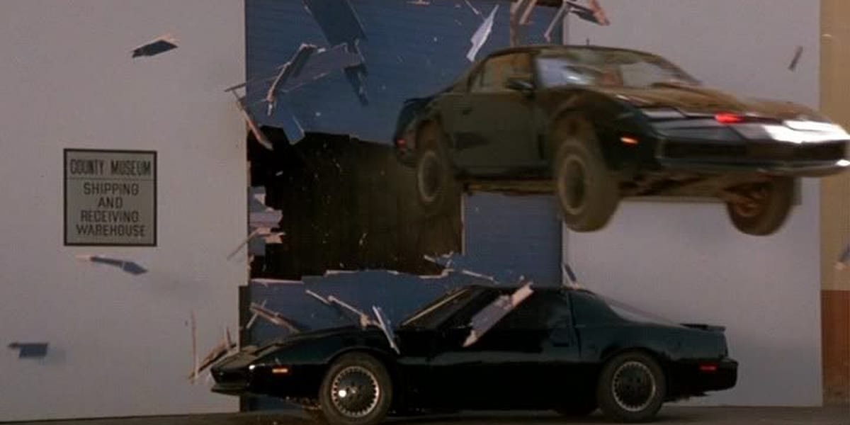 KITT jumps over another car just like him, smashing through a blue warehouse door in Knight Rider