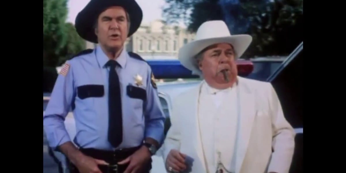 Rosco with his hands on his belt, mouth open as he speaks, with Boss standing next to him, a cigar in his mouth in Dukes of Hazzard