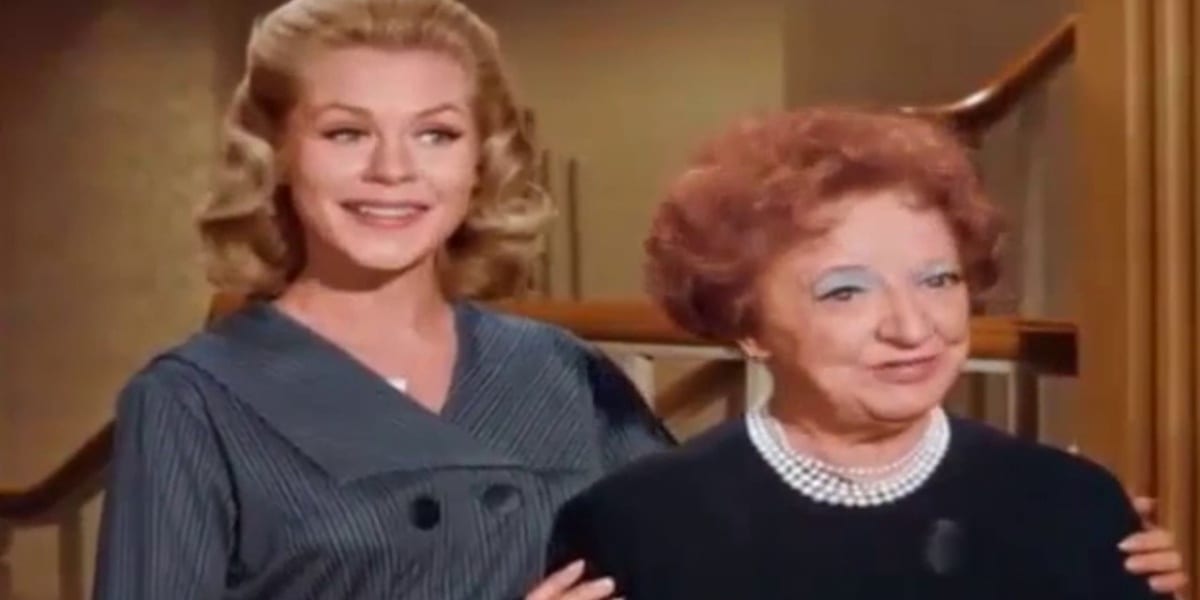 Samantha and Clara in Bewitched, Samantha smiling as she has her hands on either side of Clara's shoulders as she introduces her, Clara looking shy 
