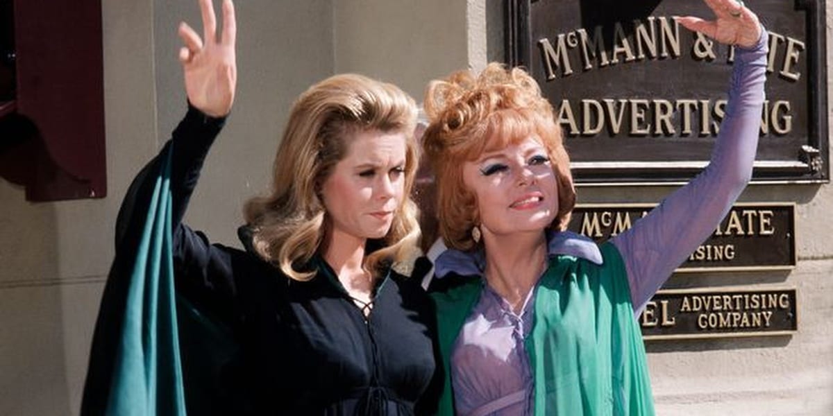 Samantha and Endora in Bewitched, Sam looking towards the ground and waving, Endora smiling and looking up, also waving, Sam wearing a black dress and cape with Endora wearing a purple and green dress
